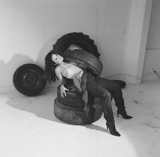 Monochrome portrait of a woman sitting on tires in a studio setting, wearing jeans and high heels, with various tires around her.