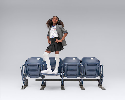 Young girl in fashionable outfit, blazer, skirt, and knee socks, standing on stadium seats, posing confidently in a studio setting.
