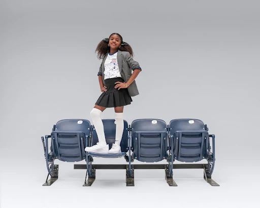 Young girl in fashionable outfit, blazer, skirt, and knee socks, standing on stadium seats, posing confidently in a studio setting.