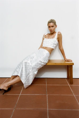 A woman in a white textured two-piece outfit leans against a wooden bench, posing in a brightly lit room with terracotta tile flooring.