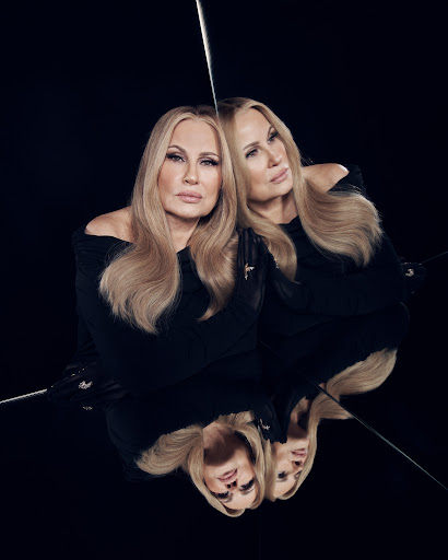 Portrait of Jennifer Coolidge reflecting in a mirror, wearing a black dress, with blond hair, against a dark background.