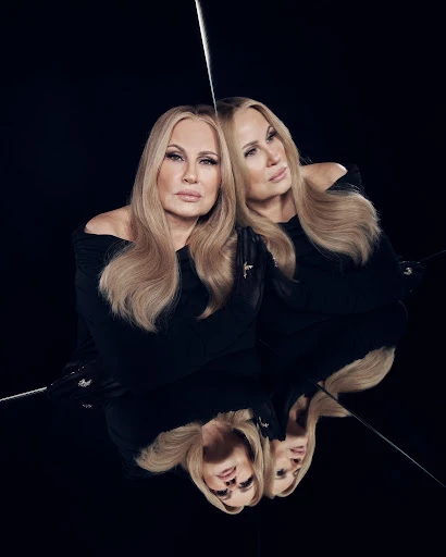 Portrait of Jennifer Coolidge reflecting in a mirror, wearing a black dress, with blond hair, against a dark background.