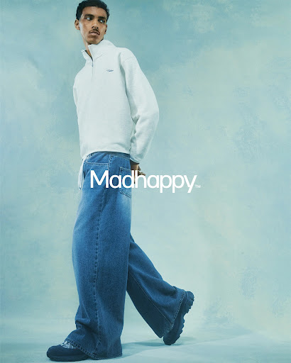 Young adult poses in a studio wearing a white zip-up sweatshirt and wide-leg denim jeans with chunky platform shoes against a blue backdrop.