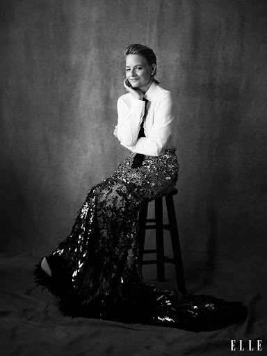 Black and white portrait of an adult female sitting on a stool, wearing a button-down shirt and long sequined skirt against a grey backdrop.