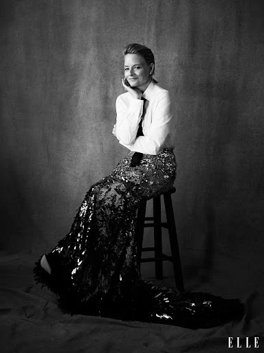 Black and white portrait of an adult female sitting on a stool, wearing a button-down shirt and long sequined skirt against a grey backdrop.