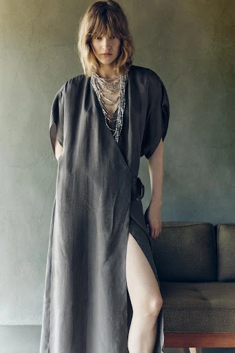 Woman wearing a wrap dress and layered necklaces, posing in front of a textured wall and a dark couch, in soft, diffused lighting.