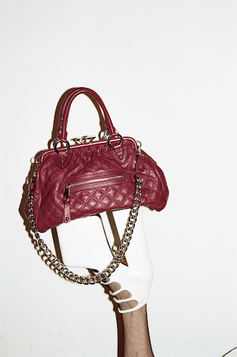 Burgundy Marc Jacobs handbag with silver chain accent displayed against a white background held by a hand wearing a white glove and fishnet sleeve.