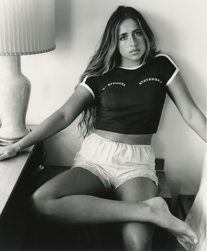 Portrait of a young woman seated indoors wearing a dark t-shirt and light shorts beside a lamp with a pleated shade.