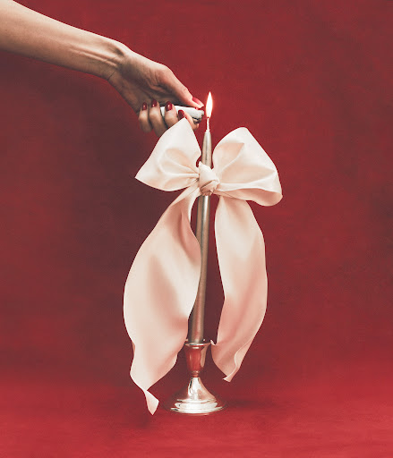 Hand lights an elegant silver candle with a large white bow against a red background, creating a sophisticated and festive scene.