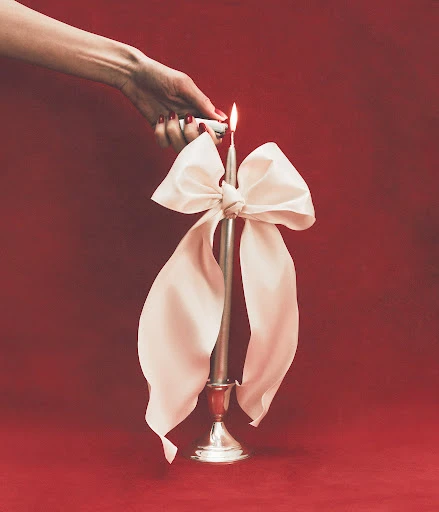 Hand lights an elegant silver candle with a large white bow against a red background, creating a sophisticated and festive scene.