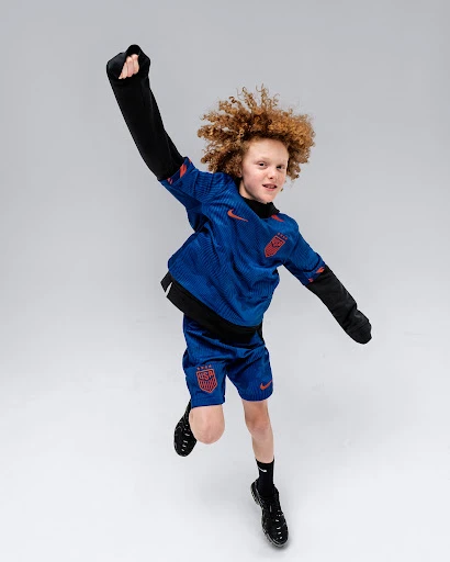 Full-length shot of a cheerful child in mid-air, wearing a blue and red athletic outfit against a bright grey background.