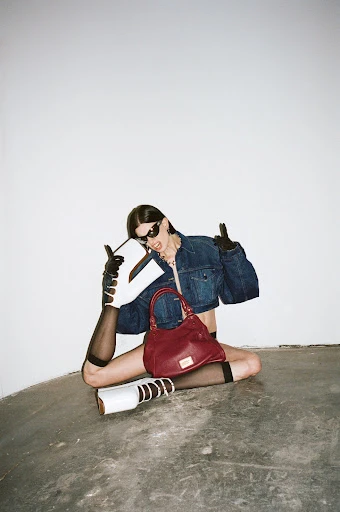 Stylish woman in denim jacket posing with a burgundy purse, white boots, and sunglasses against a white wall in a studio.