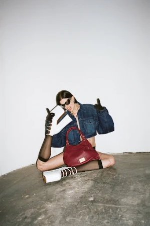 Stylish woman in denim jacket posing with a burgundy purse, white boots, and sunglasses against a white wall in a studio.