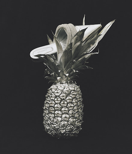 Still life of a pineapple with a slide sandal on top, set against a dark background in a visually striking, monochromatic composition.