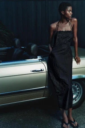 A dark-skinned woman in a black dress poses near a classic beige convertible car in an outdoor setting with dark tones.