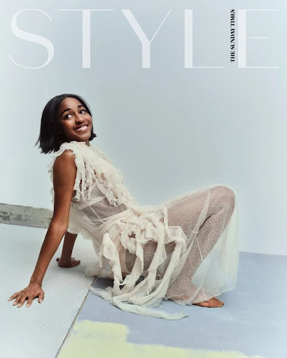 Fashion editorial shot of a smiling woman in sheer, off-white dress posing on a geometric surface in a photo studio, lit with bright, diffused light.