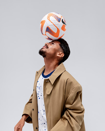 A man balances a soccer ball on his head wearing a tan coat and printed t-shirt on a neutral background, captured in a studio shot.