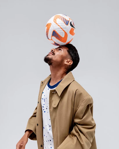 A man balances a soccer ball on his head wearing a tan coat and printed t-shirt on a neutral background, captured in a studio shot.