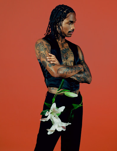 Portrait of a man with dreadlocks, tattoos, and a black vest holding a white flower against a red background in a studio shot.