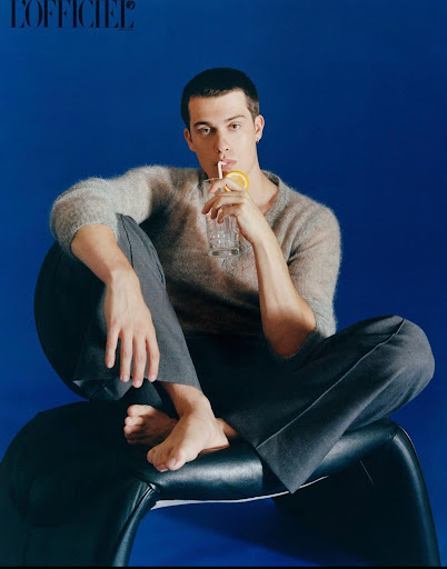 Man sits on modern black chair drinking clear beverage with straw against blue background. Gray sweater and trousers. Bare feet.