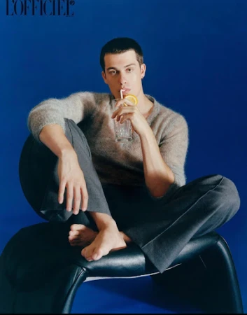 Man sits on modern black chair drinking clear beverage with straw against blue background. Gray sweater and trousers. Bare feet.