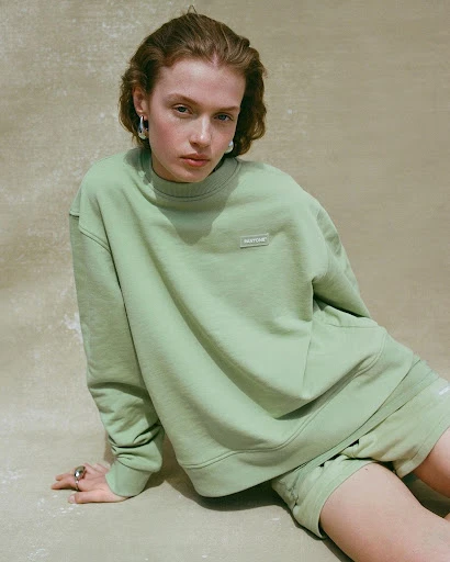 A woman poses in a light green sweatshirt and matching shorts in a studio setting, exuding a relaxed, fashionable vibe in soft lighting.