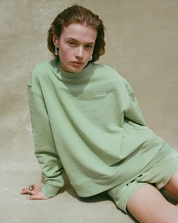 A woman poses in a light green sweatshirt and matching shorts in a studio setting, exuding a relaxed, fashionable vibe in soft lighting.