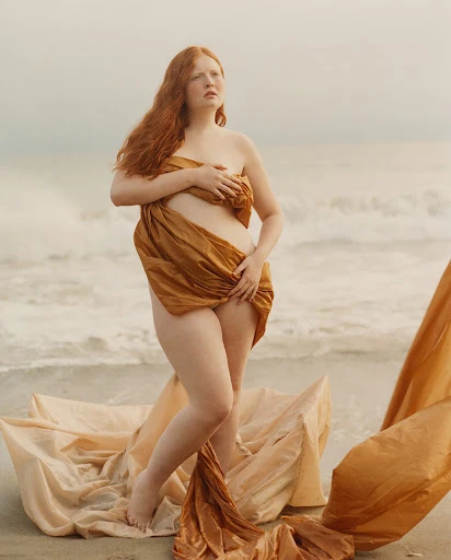 Redhead woman poses draped in gold fabric on a beach at water's edge, looking towards the horizon with a neutral expression.