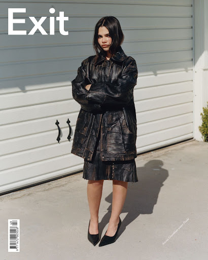 A young woman in a black leather jacket and skirt poses outdoors against a white garage door in a fashion editorial shot.
