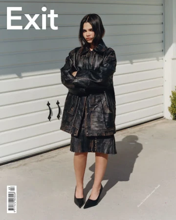 A young woman in a black leather jacket and skirt poses outdoors against a white garage door in a fashion editorial shot.