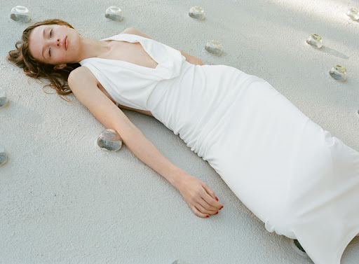 Woman in a white gown lies on a textured surface with embedded glass accents in a fashion or beauty editorial setting.