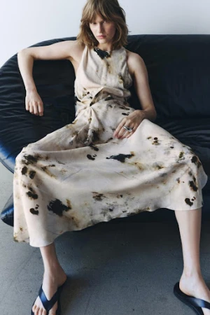 Model wearing a patterned sleeveless dress seated on a curved black leather sofa with dark flip flops.