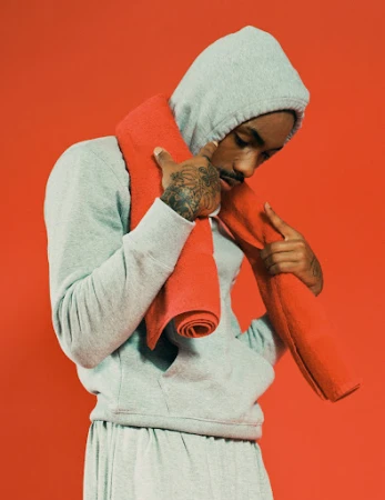 Studio shot of a male model in a grey hoodie and sweatpants holding a red scarf against a red backdrop, showcasing casual fashion.
