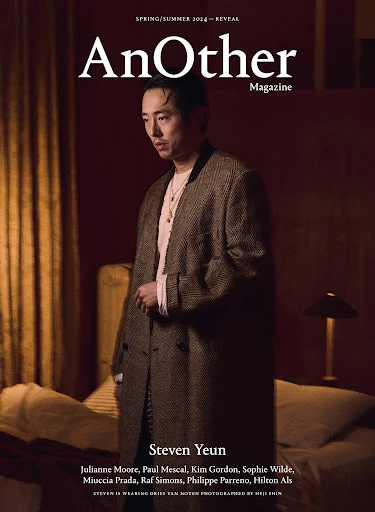 Steven Yeun on the cover of AnOther Magazine Spring/Summer 2014 issue, wearing brown overcoat indoors with lamp, bed, and red wall.