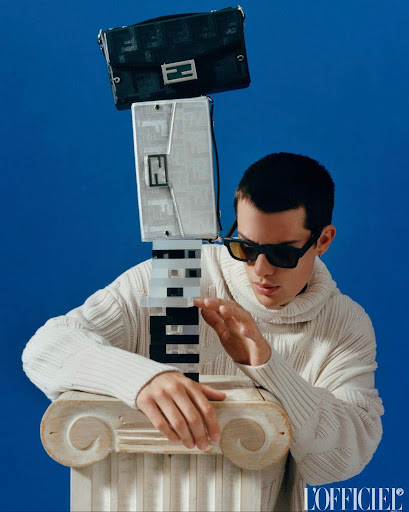 Editorial fashion shot of a man in a sweater and sunglasses beside Fendi handbag tower with a blue backdrop, clean and modern.