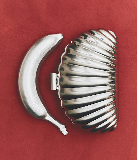 A silver-painted banana is positioned next to a silver dessert mold on a red background, creating a surreal and modern still life.