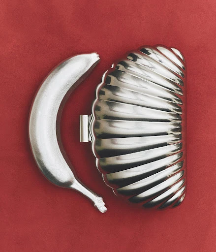 A silver-painted banana is positioned next to a silver dessert mold on a red background, creating a surreal and modern still life.
