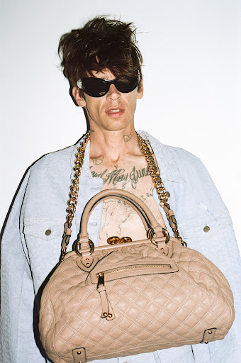 Man wearing sunglasses and light denim jacket holding a quilted beige handbag against a white background.