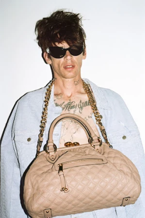 Man wearing sunglasses and light denim jacket holding a quilted beige handbag against a white background.