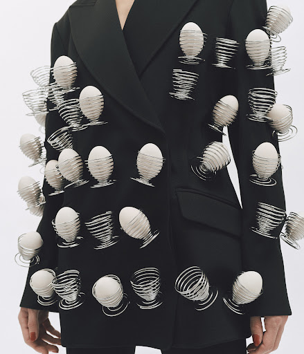 Fashion shot of a person wearing a black blazer adorned with egg cups holding white eggs, photographed on a plain white backdrop.