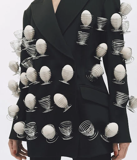 Fashion shot of a person wearing a black blazer adorned with egg cups holding white eggs, photographed on a plain white backdrop.