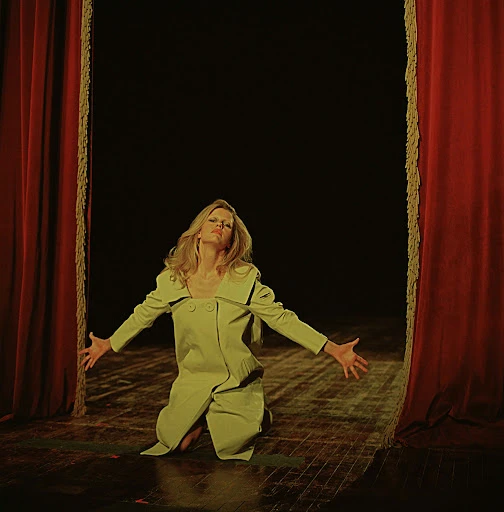Woman in yellow outfit kneeling on stage with red curtains and dark backdrop, arms outstretched in performance or expression.