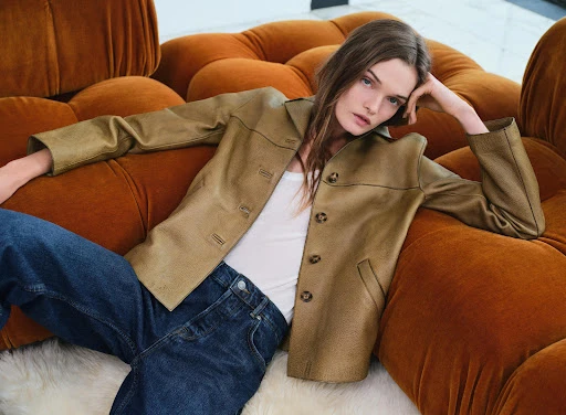 Indoor fashion shot: a model poses on an orange sofa wearing a light brown jacket, white top, and blue jeans in soft, diffused light.