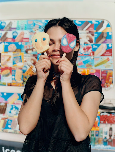 A young woman with dark hair poses outdoors, holding two ice cream pops shaped like cartoon characters in front of her face.