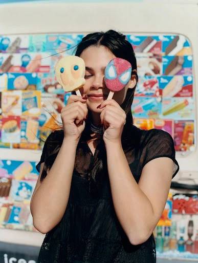 A young woman with dark hair poses outdoors, holding two ice cream pops shaped like cartoon characters in front of her face.