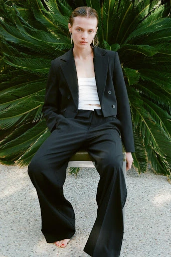 A woman in a black suit poses while sitting on a stool against a background of lush green foliage, showcasing a blend of fashion and nature.