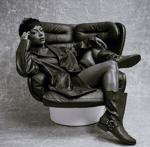 Portrait of a woman in black leather jacket, shorts, and boots sitting in an armchair against a grey studio background.
