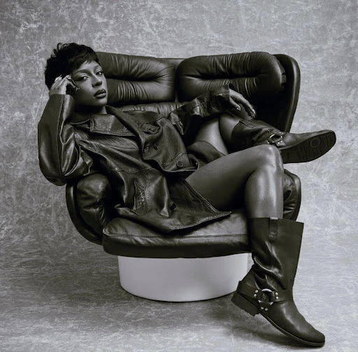 Portrait of a woman in black leather jacket, shorts, and boots sitting in an armchair against a grey studio background.