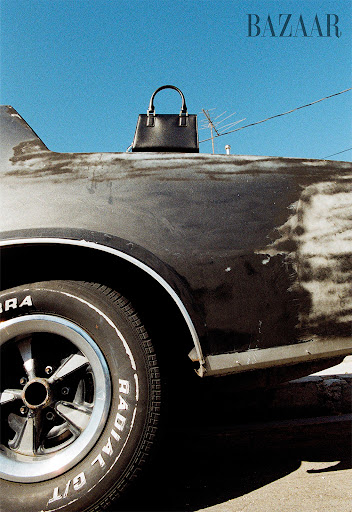 Black leather handbag on vintage car against a blue sky featuring a car tire and the word 'BAZAAR' in the upper corner.