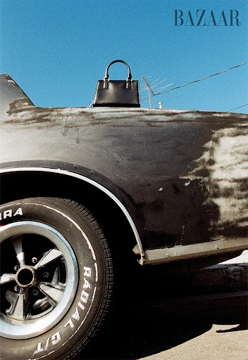 Black leather handbag on vintage car against a blue sky featuring a car tire and the word 'BAZAAR' in the upper corner.